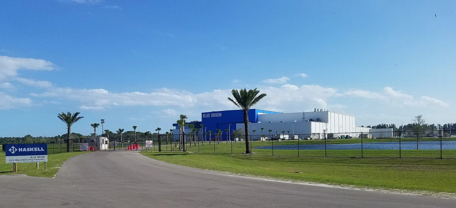 Blue Origin's large blue and white manufacturing building behind a fence, with palm trees, a pond and an entrance sign reading Blue Origin OLS Manufacturing Complex