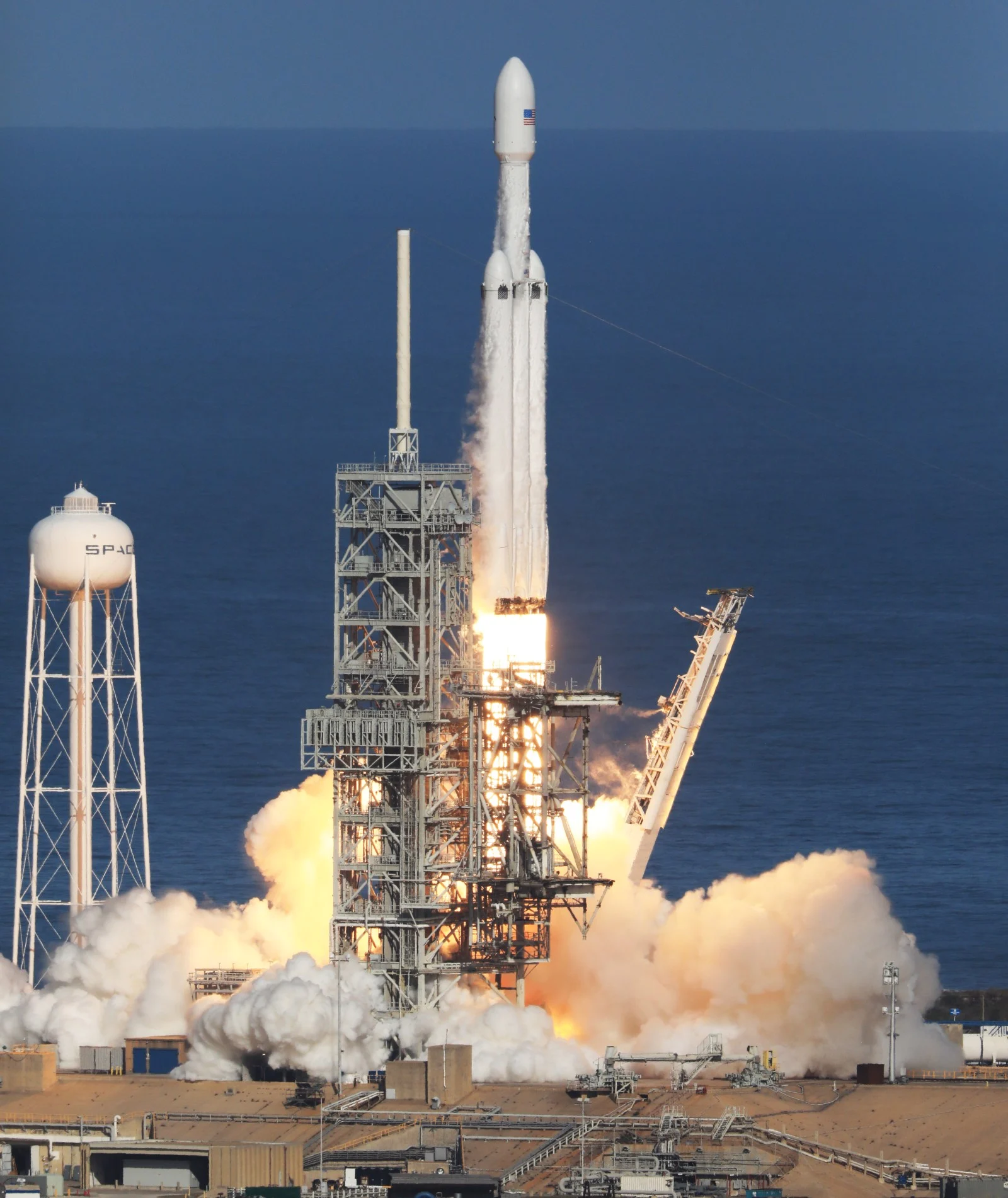 A Falcon Heavy rocket lifts off beside its launch tower, above bright exhaust and clouds of smoke.