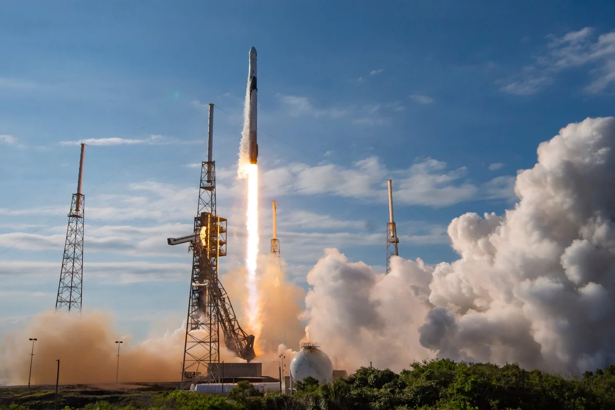 A Falcon 9 rocket rises above a launch tower with a bright exhaust plume and white clouds spreading across the pad.