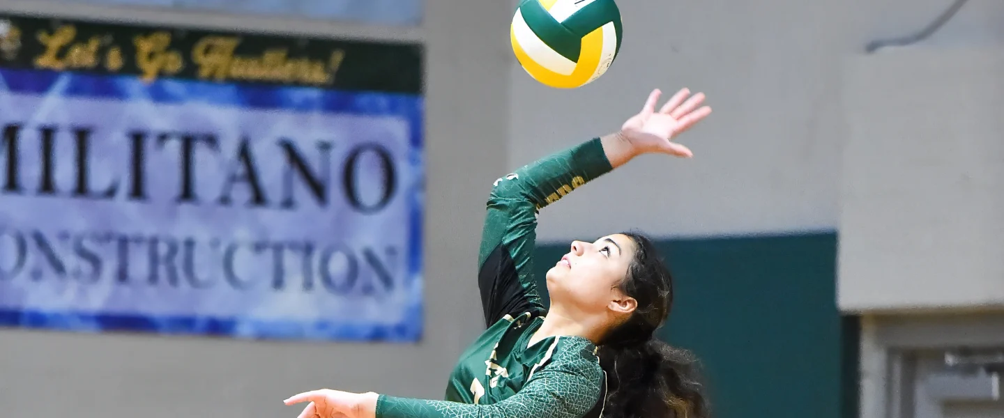 A volleyball player in a green uniform reaches toward a ball above her head.