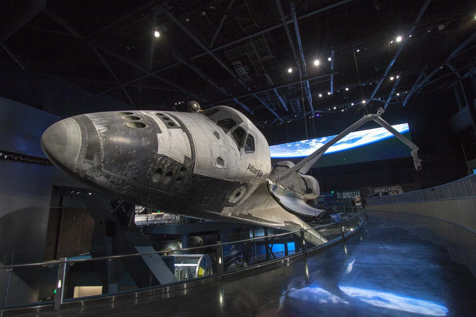 Space shuttle Atlantis displayed at an angle indoors, with its robotic arm extended and visitor walkways below.