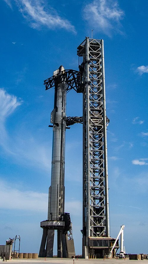 Starship and its Super Heavy booster stand beside a steel launch tower under a blue sky.