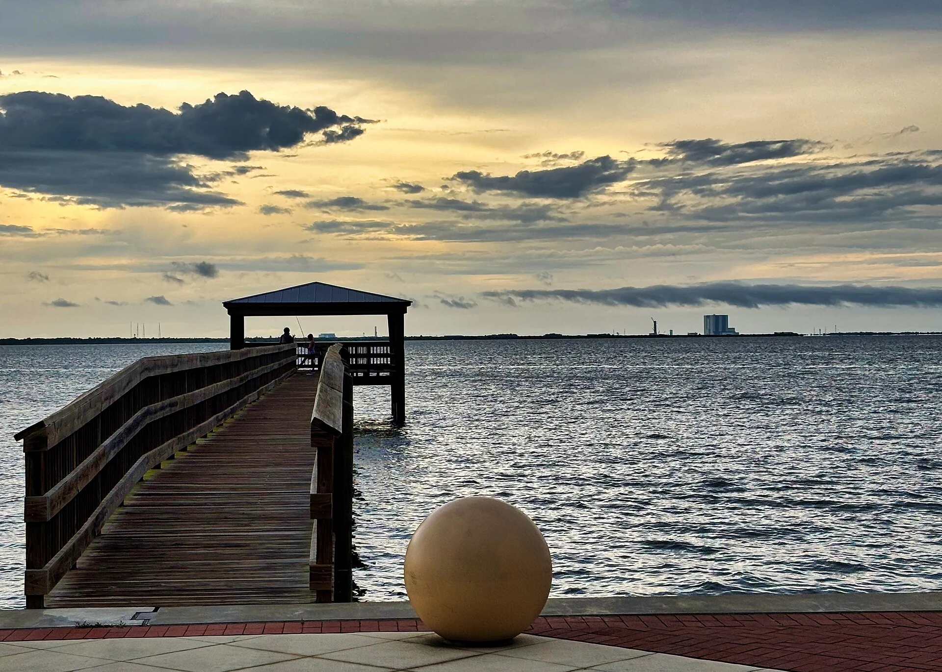 A wooden fishing pier with a covered end stretches into the Indian River under an overcast evening sky, with Kennedy Space Center's launch towers and Vehicle Assembly Building on the far shore