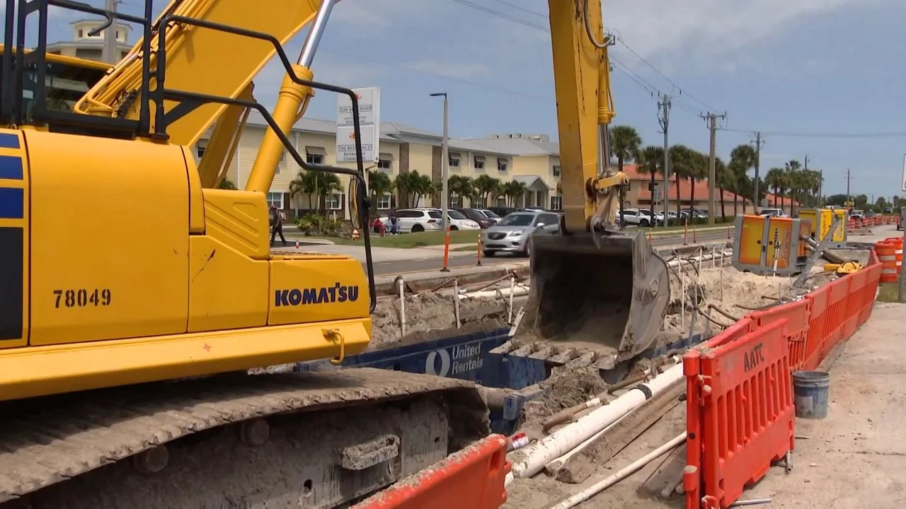 A yellow excavator beside a roadside trench and orange construction barriers