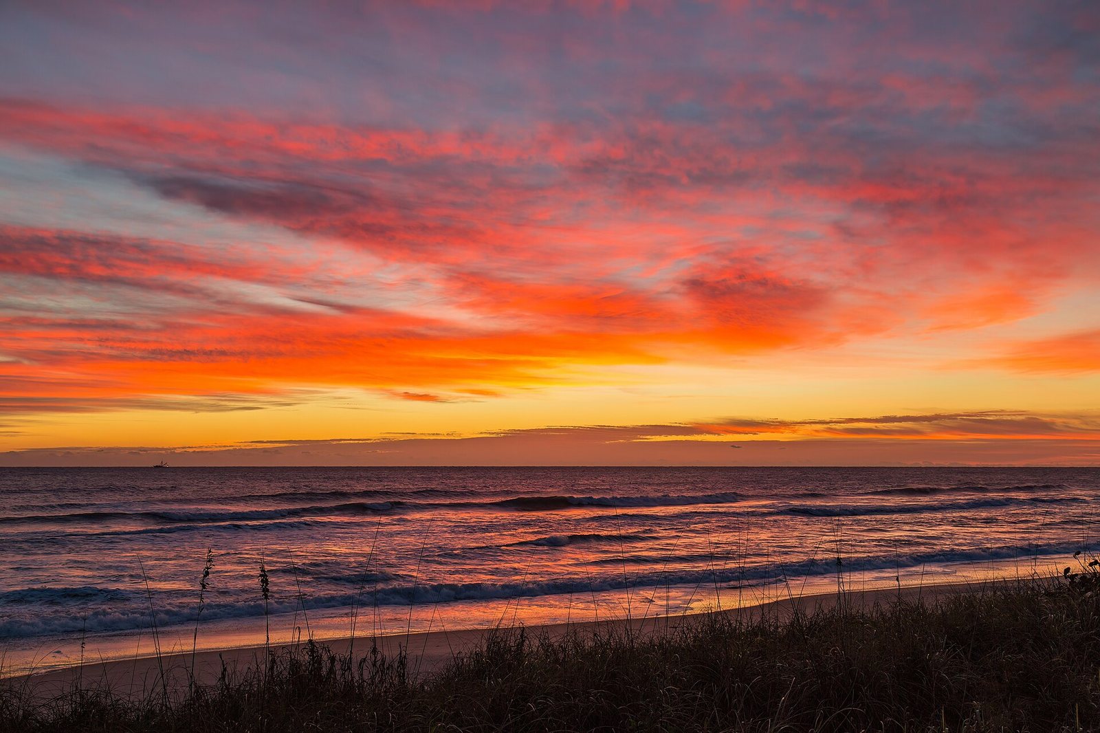 Orange and red sunrise above ocean waves, beach sand and dune grasses.